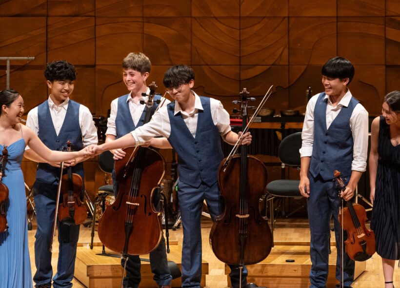 A group of young musicians joyfully playing violins and a cello together in a bright, lively setting