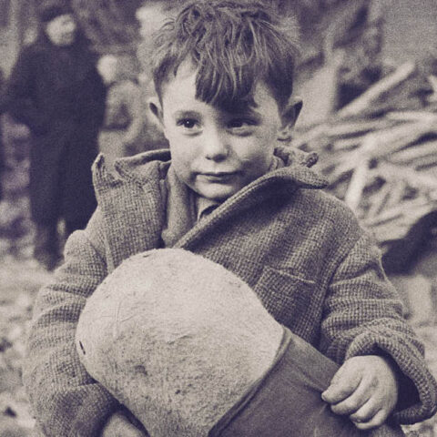 A young boy sits on rubble holding a stuffed toy, with damaged buildings and people in the background. 