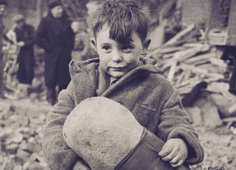 A young boy sits on rubble holding a stuffed toy, with damaged buildings and people in the background. 