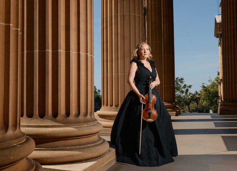 A woman in a black ball gown standing before a row of columns, with a violin in hand