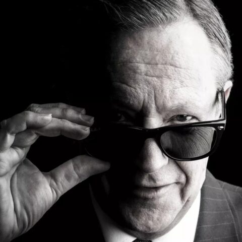 Black-and-white portrait of a suited older man adjusting dark sunglasses, staring at the camera with a confident, mysterious expression