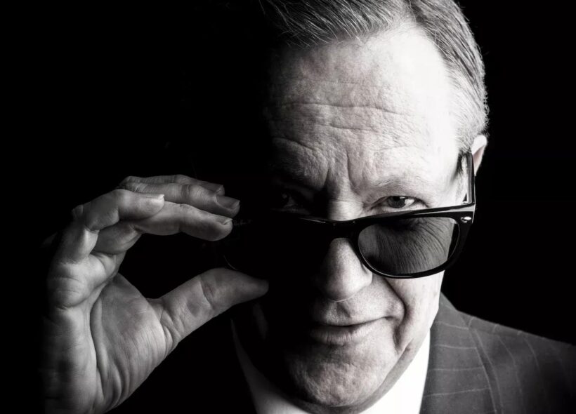 Black-and-white portrait of a suited older man adjusting dark sunglasses, staring at the camera with a confident, mysterious expression