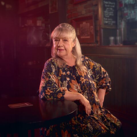 Smiling older woman in a floral dress sits at a pub table, surrounded by framed art in warm, moody lighting