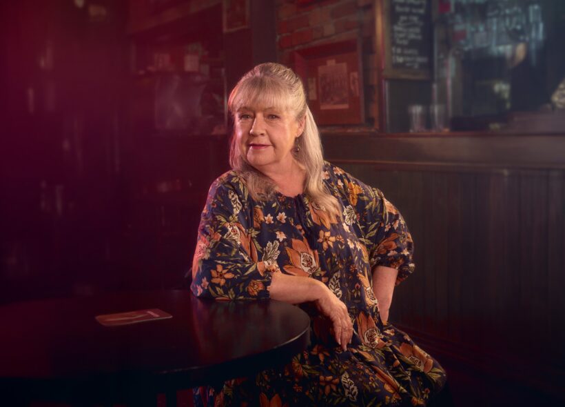 Smiling older woman in a floral dress sits at a pub table, surrounded by framed art in warm, moody lighting
