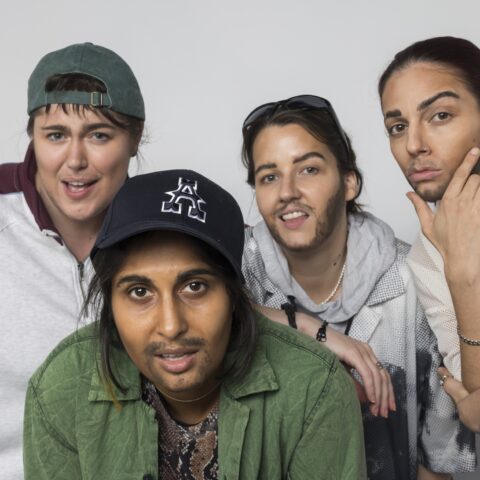 Four people pose together, each wearing street clothes with hats or sunglasses, with drawn-on facial hair, against a plain light background. 