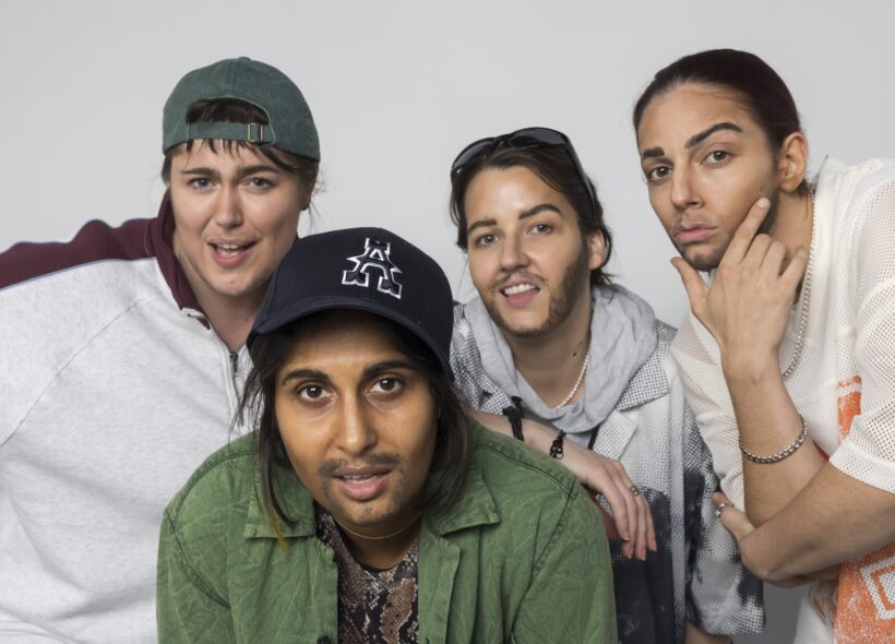 Four people pose together, each wearing street clothes with hats or sunglasses, with drawn-on facial hair, against a plain light background. 