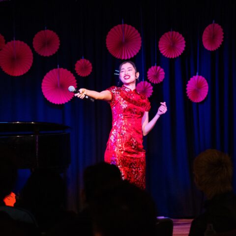 A woman in a red dress stands confidently on stage, engaging the audience with her presence. 