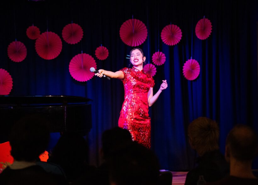 A woman in a red dress stands confidently on stage, engaging the audience with her presence. 