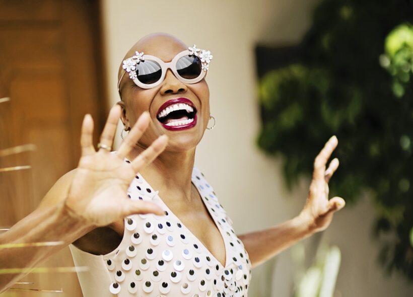 A woman poses with her hands up, smiling brightly, wearing bold sunglasses 