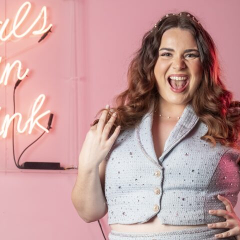 Joyful woman with wavy hair poses in a pink room, wearing a light blue outfit; neon script behind her reads “Pals in Pink.”