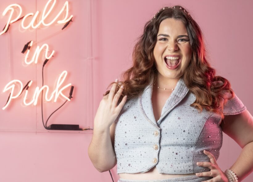 Joyful woman with wavy hair poses in a pink room, wearing a light blue outfit; neon script behind her reads “Pals in Pink.”