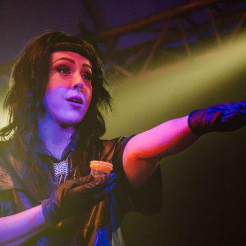 Cleo, a caucasian woman with long black hair and front bangs, is holding a rhinestoned specimen cup full of urine with her left arm outstretched. She wears a leather shirt, roman collar, and disposable gloves.