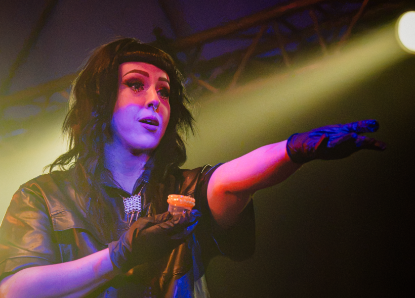 Cleo, a caucasian woman with long black hair and front bangs, is holding a rhinestoned specimen cup full of urine with her left arm outstretched. She wears a leather shirt, roman collar, and disposable gloves.