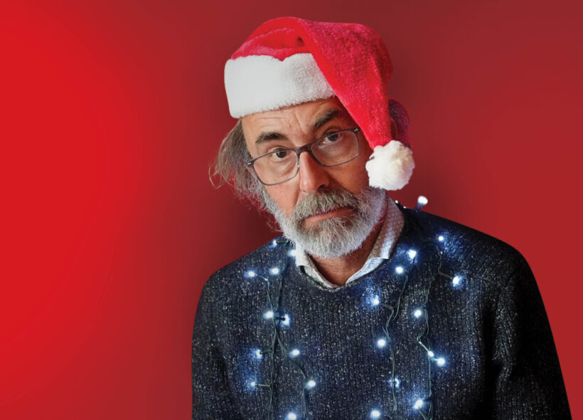 A man wearing a festive Santa hat and glasses with Christmas lights around his neck, stares unsmiling at the camera. 