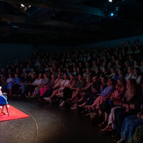 A woman speaks from a blue chair on a red rug beside flowers, facing a packed, dim theater audience in an intimate, attentive atmosphere.