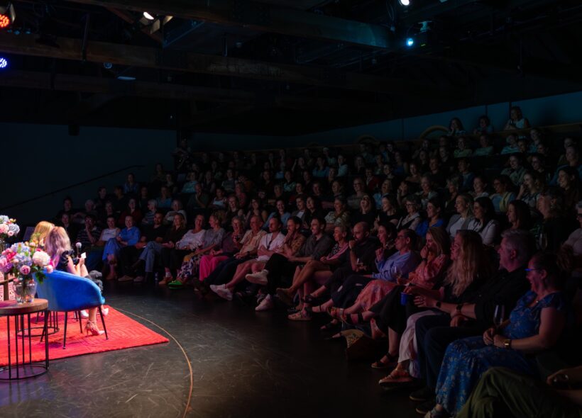 A woman speaks from a blue chair on a red rug beside flowers, facing a packed, dim theater audience in an intimate, attentive atmosphere.