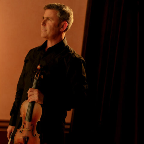 A Man in black holds a violin and stares off camera to the left