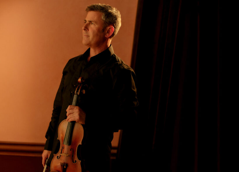 A Man in black holds a violin and stares off camera to the left