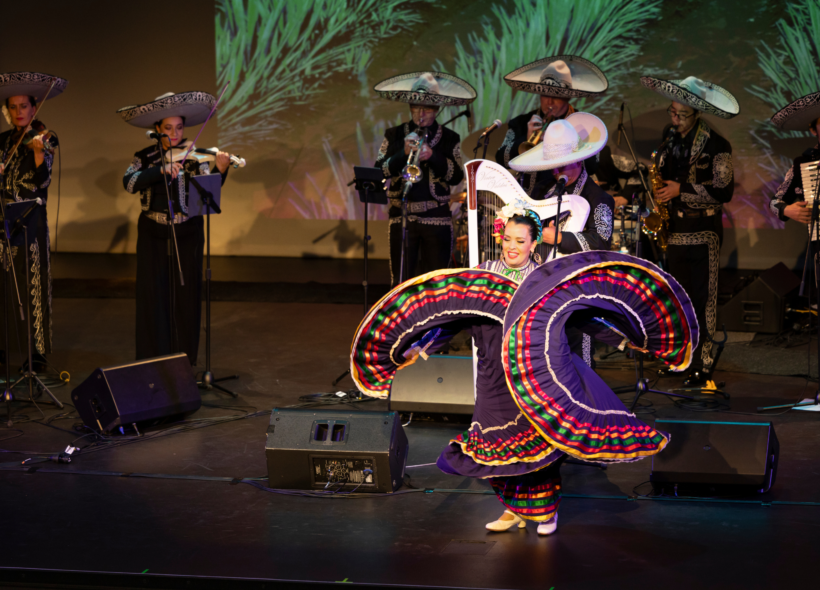 A woman dances on stage in a colourful dress, behind her a mariachi band plays 