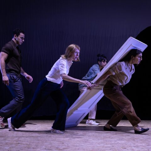 Four theater performers rush a large white door across a dim, purple-lit stage, creating a comic, urgent sense of movement.