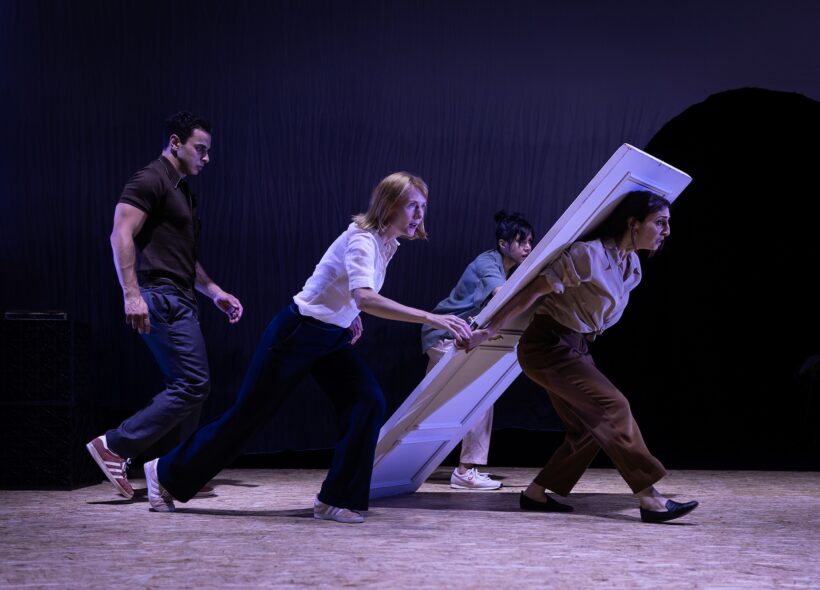 Four theater performers rush a large white door across a dim, purple-lit stage, creating a comic, urgent sense of movement.